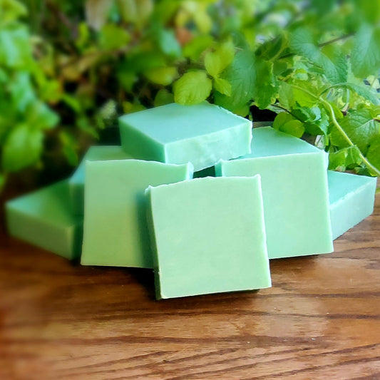 Bright Green colored square soap with natural mica handcrafted with care by Gothic & Ivy, are stacked and arranged in front of a grey planter containing three varieties of freshly growing mint.