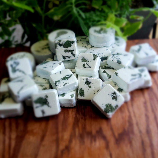 Square-shaped shower steamers colored mildly with natural green mica topped with organically grown dried spearmint from the planter behind them.