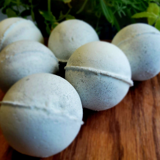 Swirling pattern of pale grey and white bath bombs, colored with natural mica handcrafted with care by Gothic & Ivy, arranged in front of a grey planter containing three varieties of freshly growing mint.