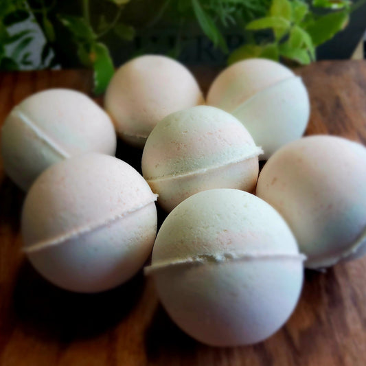 Swirling pattern of pale green and pink bath bombs, colored with natural mica handcrafted with care by Gothic & Ivy, arranged in front of a grey planter containing three varieties of freshly growing mint.