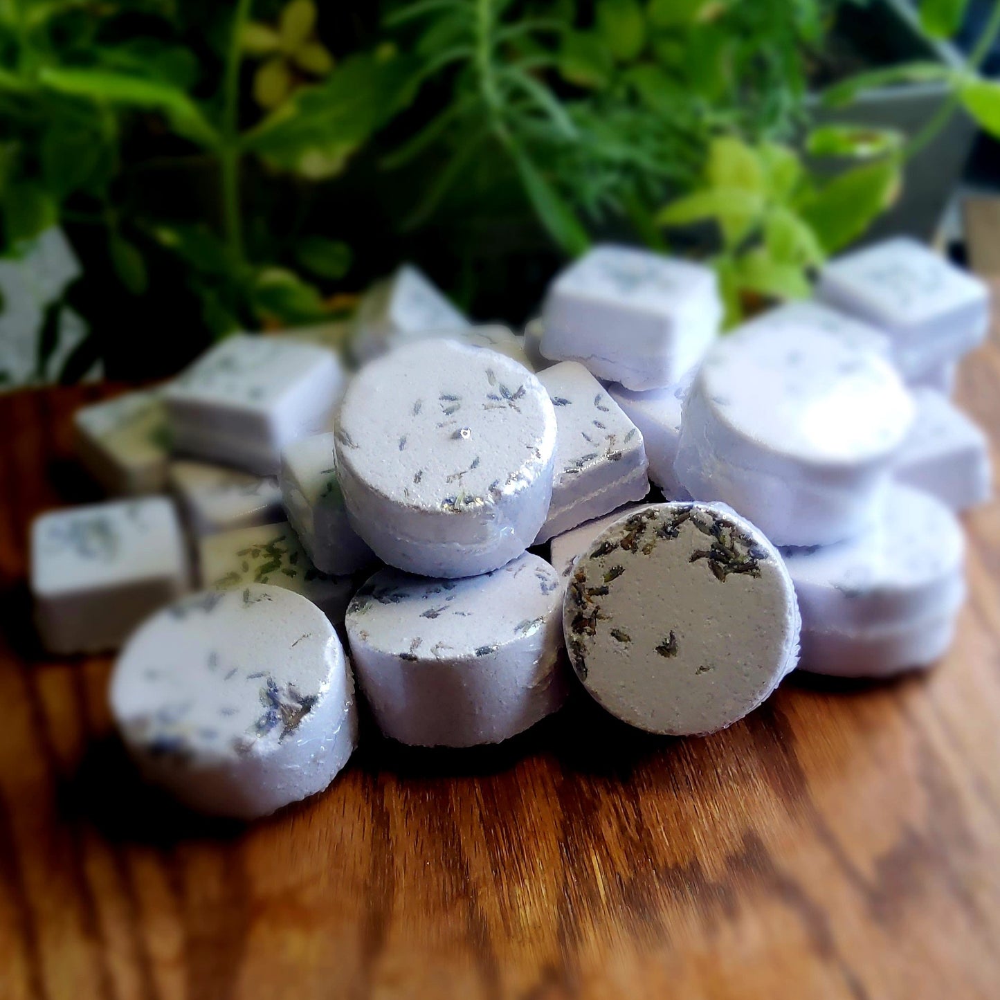 One inch round shower steamers colored pale purple mica and topped with organic lavender flower buds, handmade in small batches by Gothic & Ivy. With a grey planter behind them showcasing three different kinds of mint.