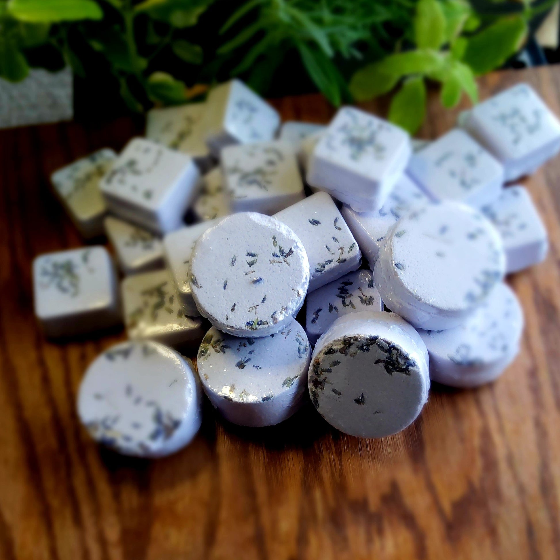 One inch round shower steamers colored pale purple mica and topped with organic lavender flower buds, handmade in small batches by Gothic & Ivy. With a grey planter behind them showcasing three different kinds of mint.