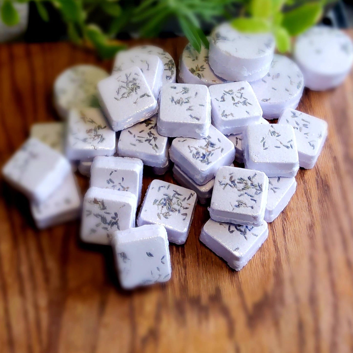 Square-shaped shower steamers colored pale purple mica and topped with organic lavender flower buds, handcrafted in small batches by Gothic & Ivy. With a grey planter behind them showcasing three different kinds of mint.