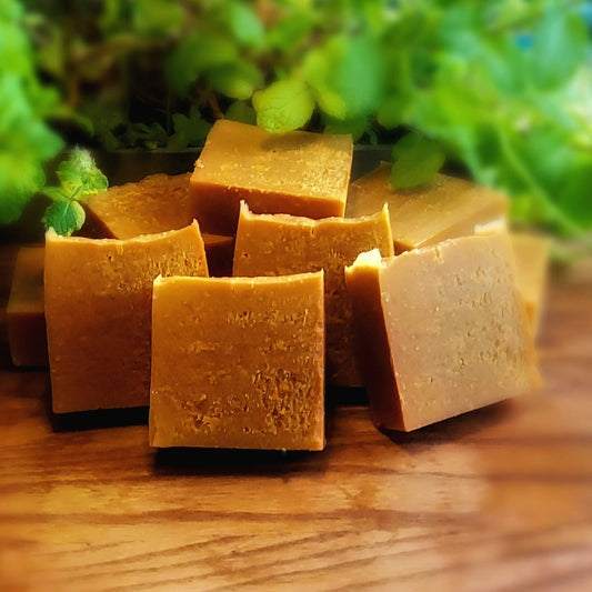 Rich, darkly orange colored square soap handcrafted with care by Gothic & Ivy, are stacked and arranged in front of a grey planter containing three varieties of freshly growing mint.